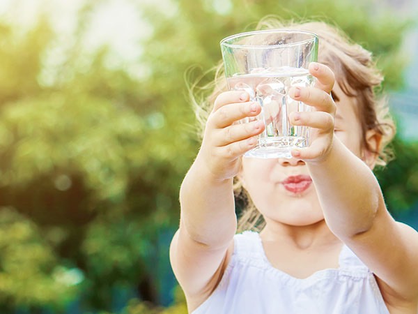 Ett barn i vit klänning håller upp ett glas klart dricksvatten med båda händerna i solen. Bakgrunden är grön och suddig, vilket förstärker känslan av en somrig dag utomhus och betonar vattenglaset i förgrunden.