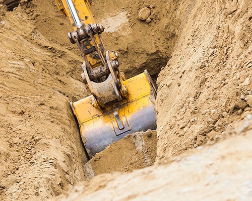 En gul grävmaskin i arbete, sänker skopan i en smal jordtrench. Omgiven av brun jord, flyttar skopan material för en byggprojekt. Detaljer i maskinen visar robust konstruktion och mekanik, typisk för tunga anläggningsmaskiner.