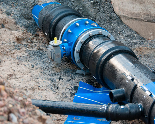 En stor, blå vattenledning med metallflänsar och en ventil ligger delvis begravd i jord. Intill ledningen syns en svart mindre rörledning. Omgivningen består av jord och sten, vilket antyder pågående arbete med rörläggning.