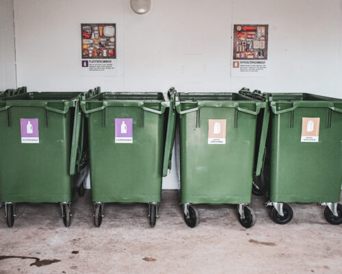 Gröna soptunnor med hjul står uppradade mot en vit vägg inomhus. De är märkta för återvinning av plast- och pappersförpackningar med färgglada etiketter. Informationsskyltar syns ovanför behållarna.