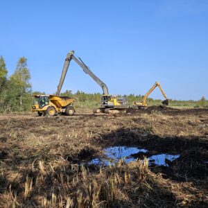 http://Två%20grävmaskiner%20arbetar%20på%20en%20öppen,%20lerig%20åker.%20Den%20ena%20maskinen%20lyfter%20jord%20till%20en%20lastbil%20medan%20den%20andra%20gräver%20i%20marken.%20I%20bakgrunden%20syns%20träd%20och%20en%20klar%20blå%20himmel.
