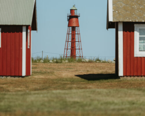 Röd fyr står mellan två rödvita stugor på en grönskande äng under en klarblå himmel. En fågel svävar över den vänstra stugan. Marken är torrare i förgrunden med gröna buskar i bakgrunden.