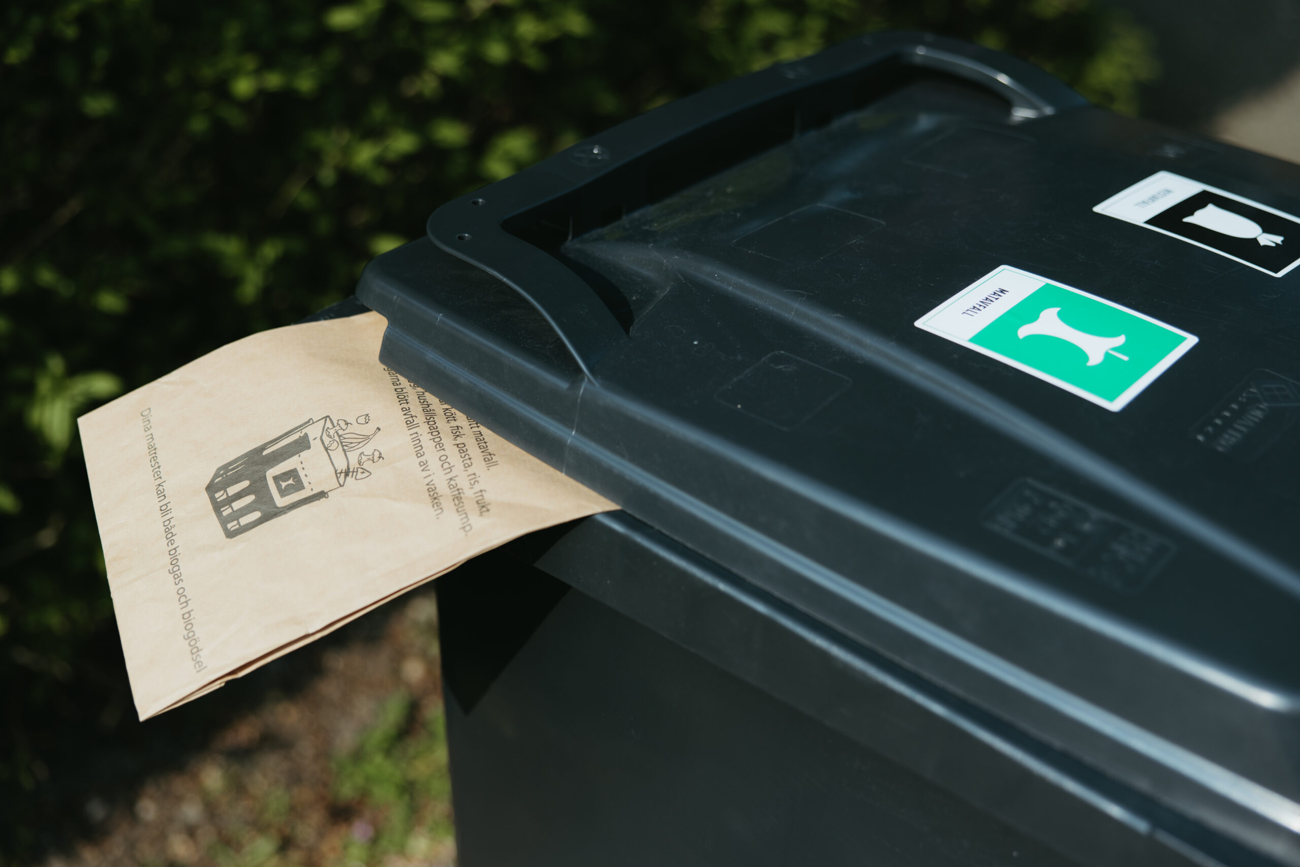 En papperspåse klämd i locket på ett svart sopkärl.