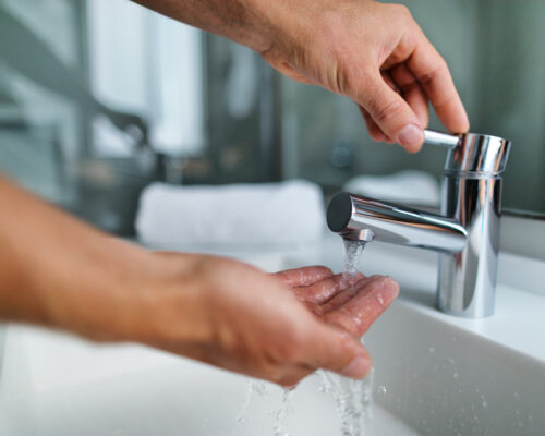 Man som tvättar händerna i badrumshandfatet hemma, kontrollerar temperaturen och rör vid rinnande vatten med handen. Närbild på fingrar under varmt vatten från en kran.