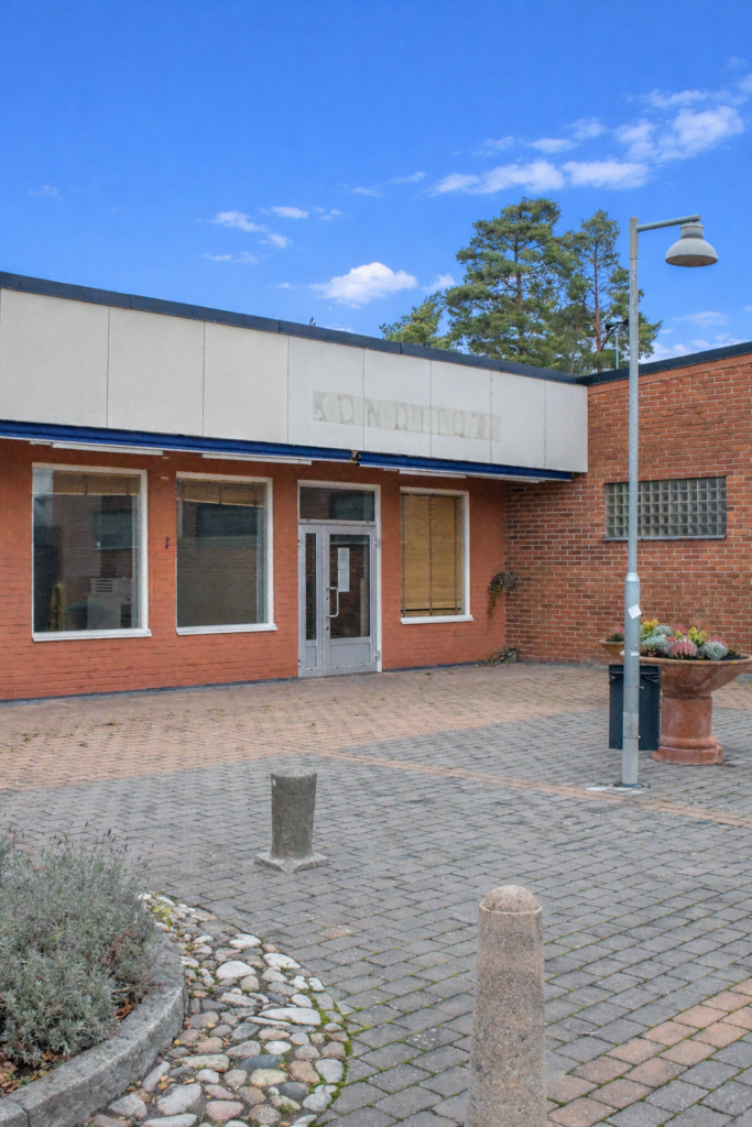 Utsidan av en företagslokal med stora butiksfönstel och glasad entrédörr. Vackert belägen stenläggning utanför.