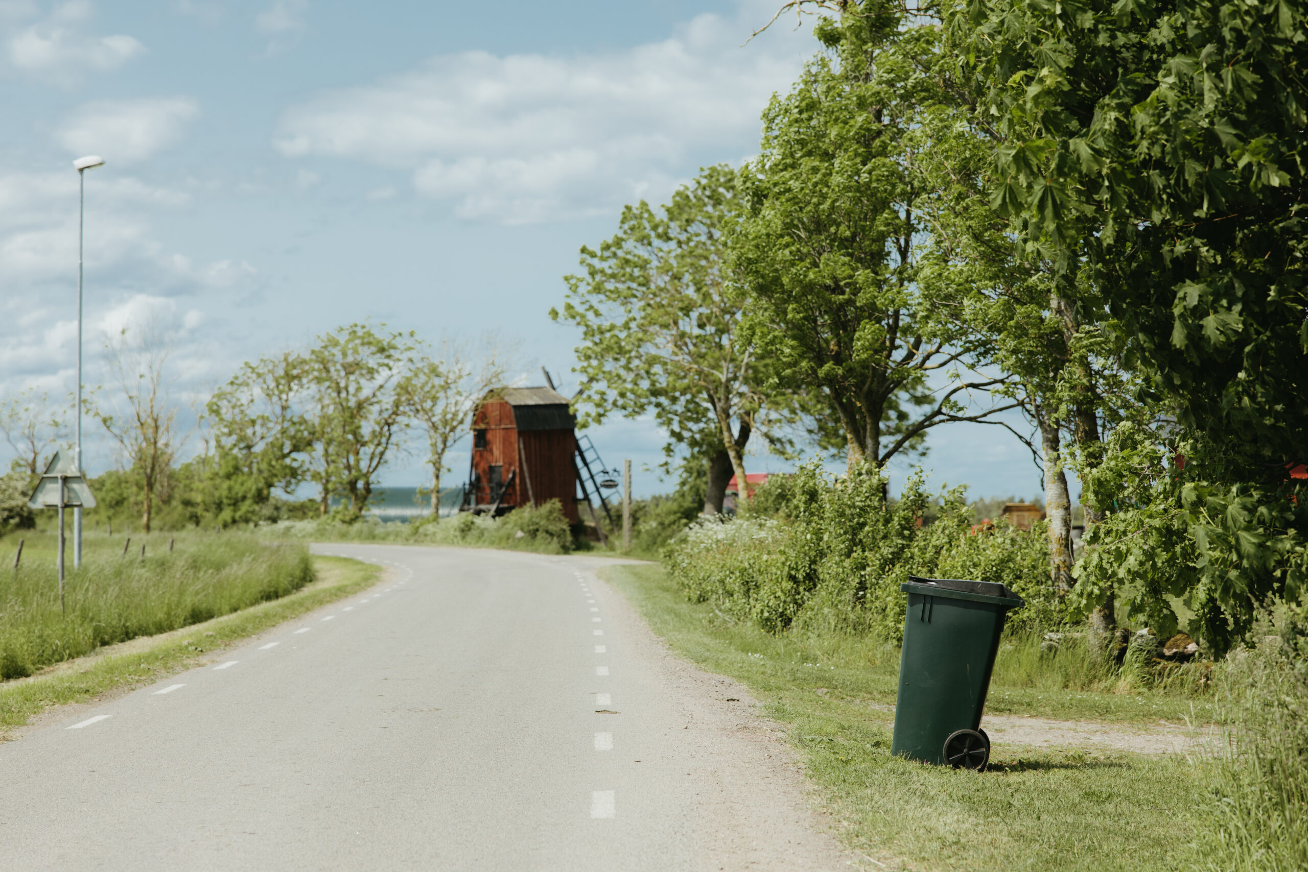 Ett grönt sopkärl stående på sidan av en väg, en röd väderkvarn syns bland träden i bakgrunden