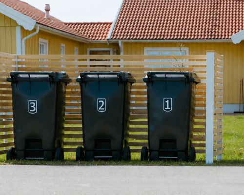 Tre svarta sopkärl står samlade i ett stilrent sopkärlsskydd av trä med horisontella ribbor framför ett gult hus med röda takpannor.